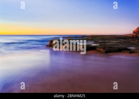 Glatte verschwommene Oberfläche des pazifischen Ozeans über dem Sandstrand von TUrimetta, der sich um Sandsteinfelsen mit Algen erstreckt. Farbenfrohe Sonnenaufgangs-Seenlandschaft. Stockfoto