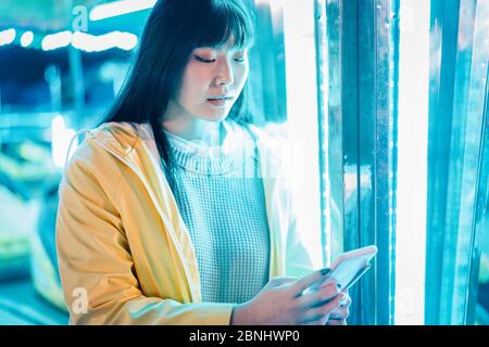 Glücklich asiatische Mädchen mit Smartphone im Freizeitpark - Junge trendige Frau mit Spaß mit Technik-Trends mit Neons in Hintergründen - Tech und Jugend c Stockfoto