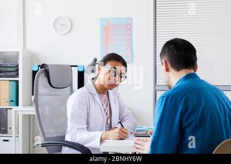 Junge Ärztin sitzt am Tisch und macht Notizen und hört dem Patienten während seines Besuchs im Büro zu Stockfoto