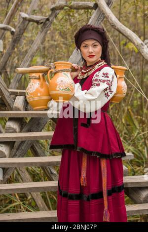 Eine Frau in traditioneller Kleidung trägt Wasser in alten Fässern. Alte Krüge werden von einem Mädchen in einem stilisierten Anzug getragen. Schöne, originelle Kleidung der Stockfoto