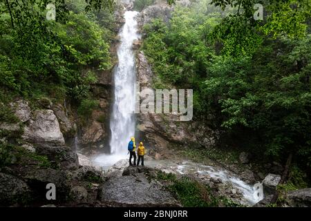 Rückansicht von Mutter und Sohn auf einem Felsen durch Wasserfall im Wald stehend Stockfoto