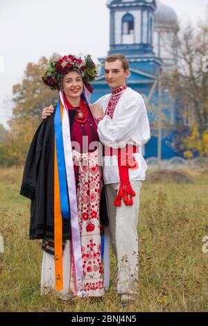 Mädchen und Kerl in Retro-Kostümen auf der Straße im alten Dorf. Retro-Inszenierung eines alten Ritus. Schöner Kranz auf einem Mädchen. Der Kerl umarmt das Mädchen Stockfoto