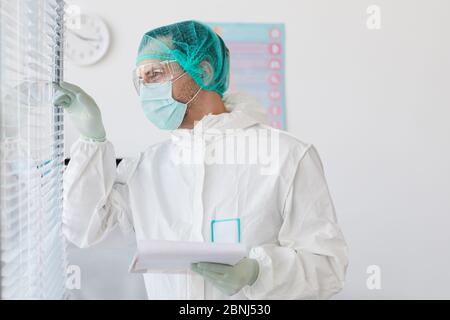 Der männliche Arzt trägt Schutzkleidung und schaut durch die Rollläden, während er im Krankenhaus mit Dokumenten arbeitet Stockfoto