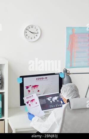 Rückansicht des Arztes in Schutzkleidung, der im Büro arbeitet, untersucht sie die Covid-Statistiken für die letzte Periode Stockfoto