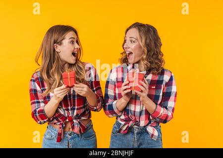 Bild von zwei jungen schönen Mädchen tragen karierten Hemden lächelnd und halten Geschenkboxen isoliert über gelbem Hintergrund Stockfoto