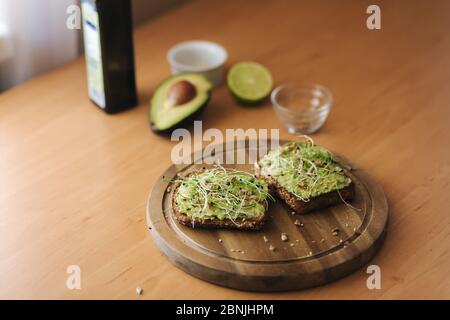 Veganer Toast mit Guacamole auf Holzbrett. Roggen-Toastbrot mit Avocado darauf. Hintergrund der Zutaten. Avocado, Limette, Olivenöl Stockfoto