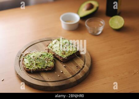 Veganer Toast mit Guacamole auf Holzbrett. Roggen-Toastbrot mit Avocado darauf. Hintergrund der Zutaten. Avocado, Limette, Olivenöl Stockfoto