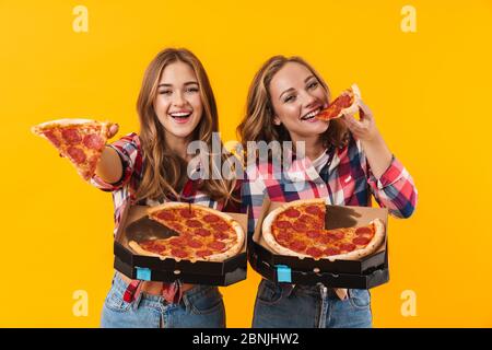 Bild von zwei jungen schönen Mädchen tragen karierten Hemden essen Pizza isoliert über gelbem Hintergrund Stockfoto