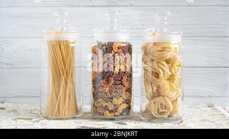 Drei Glasflaschen mit verschiedenen Nudelsorten. Pasta zu Hause auf weißem Holzregal Spaghetti creste di gallo fettuccine Stockfoto