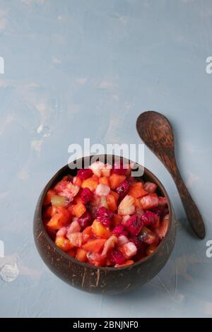 Tropischer Obstsalat in Kokosnussschale auf hellblauem Hintergrund, Nahaufnahme, Vertikales Format, Platz für Text Stockfoto