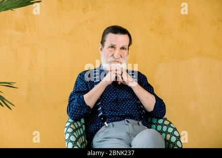 Nachdenklicher reifer bärtiger Mann, der in einem Stuhl sitzt und die Kamera anschaut. Senior männlich in stilvoller Kleidung träumen von etwas, in Gedanken verloren. Studioaufnahme Stockfoto