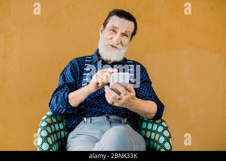 Porträt von angenehmen Senior lächelnd bärtigen Mann, trägt stilvolle Hemd und Hose, sitzt auf dem gelben Hintergrund und mit dem Telefon Apps oder soziale Stockfoto