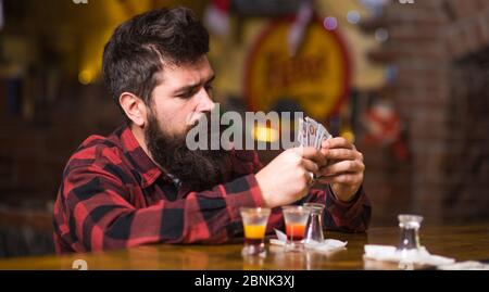 Get Drunk Concept. Mann mit betrunkenem Gesicht sitzen alleine an der Theke. Hipster hält Geld, zählen Bargeld, um mehr Alkohol zu kaufen. Guy verbringen Freizeit in Bar trinken Cocktail, defokussed Hintergrund. Stockfoto