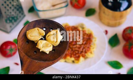 Drei Cappelletti in einem Holzlöffel und im Hintergrund ein Tisch mit typisch italienischem Essen Stockfoto