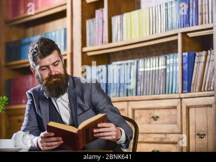 Altmodischer Mann in der Nähe der Teekanne. Aristokratische Traditionen Konzept. Aristokrat auf lächelndem Gesicht liest Buch. Mann in klassischen Anzug sitzt in Vintage-Interieur, Bibliothek, Bücherregale auf Hintergrund. Stockfoto