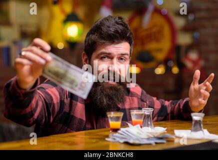 Hipster hält Geld, Bargeld, um mehr Alkohol zu kaufen. Mann mit betrunkenem Gesicht sitzen alleine an der Theke. Guy verbringen Freizeit in Bar trinken Cocktail, defokussed Hintergrund. Get Drunk Concept. Stockfoto