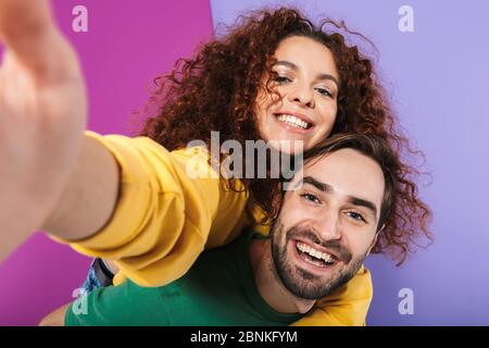 Porträt von fröhlichen lächelnden Paar Mann und Frau in bunten Kleidung, die Selfie-Foto während der Huckepack Fahrt isoliert über lila Hintergrund Stockfoto
