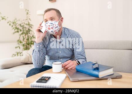 Zwangloser Geschäftsmann, der zu Hause arbeitet. Büroarbeiter Kerl in medizinischen Maske auf seinem Gesicht mit Handy. Stockfoto