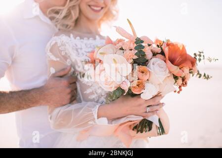 Nahaufnahme des Bouquets einer Braut aus Amaryllis, Anthurium, Rosen, Nelken, Eukalyptus in Pfirsich-Schattierungen. Die Braut in einem weißen Spitzenkleid Stockfoto