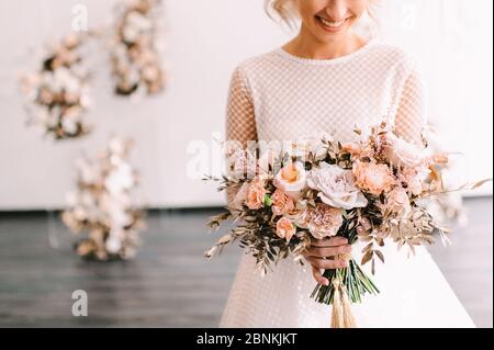 Nahaufnahme des Straußes einer Braut aus Pfirsich-Rosen, Nelken, goldenen Eukalyptusblättern. Die Braut in einem weißen Kleid mit langen Ärmeln hält einen Strauß in Stockfoto