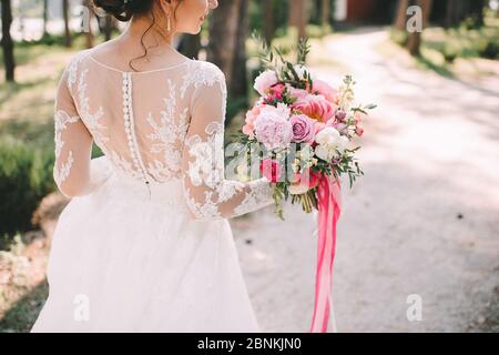 Nahaufnahme des Bouquets einer Braut aus Pfingstrosen, Rosen, Eukalyptus in weiß-rosa Schattierungen, die mit rosa Bändern gebunden sind. Die Braut hält einen Strauß in der Hand in einem Wh Stockfoto
