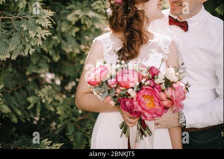 Brautstrauß Nahaufnahme, der Strauß besteht aus roten Pfingstrosen, roten und weißen Rosen. Die Braut hält den Strauß neben dem Bräutigam, in einem weißen gekleidet Stockfoto