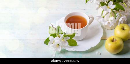 Weiße Tasse Tee auf geformter Untertasse mit blühenden Ästen von Apfelbäumen und zwei gelben Äpfeln auf Holzhintergrund mit Bokeh. Apple Urlaub. Apple Day. Stockfoto