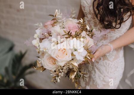 Nahaufnahme des Straußes einer Braut aus Pfirsich-Rosen, Nelken, goldenen Eukalyptusblättern. Die Braut in einem weißen Kleid mit langen Ärmeln hält einen Strauß in Stockfoto