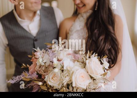 Nahaufnahme des Straußes einer Braut aus Pfirsich-Rosen, Nelken, goldenen Eukalyptusblättern. Die Braut in einem weißen Kleid mit langen Ärmeln hält einen Strauß in Stockfoto