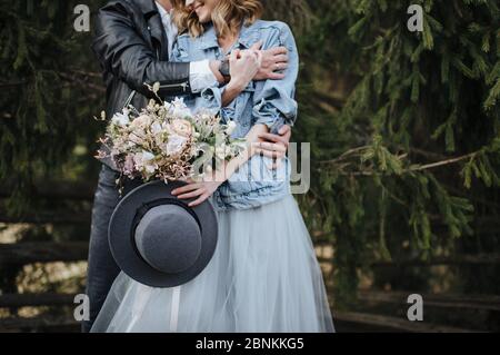 Die Braut in einer Denim-Jacke hält einen Hut und einen Strauß in Weiß- und Lila-Tönen. Bräutigam umarmt sie von hinten Stockfoto