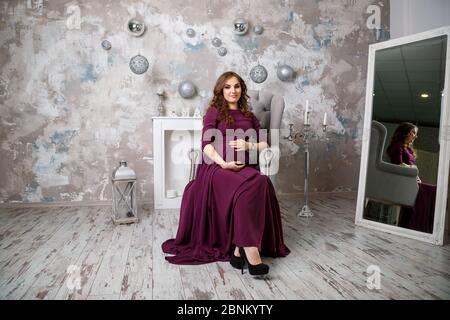 Schwangere Brünette Mädchen sitzt auf der Sessel, schaut in die Ferne, glückliche Mutterschaft. Graue Betonwand und graue Polstersessel. Stockfoto