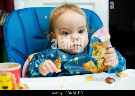 Ein kleines Kind sitzt in einem Futterstuhl und isst zum ersten Mal Brot. Gesicht in Krümel Stockfoto