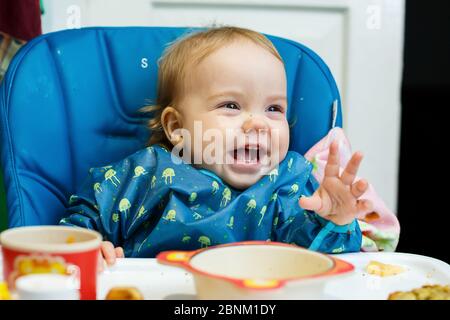 Ein kleines Kind sitzt in einem Futterstuhl und isst zum ersten Mal Brot. Gesicht in Krümel Stockfoto