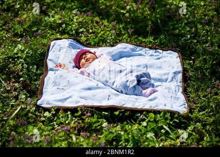 Ein Foto-Porträt eines Babys in einer roten Mütze liegt auf einem weißen Karomuschel auf grünem Gras. Der Frühling ist auf der Straße, die Sonne scheint auf das Kind Stockfoto