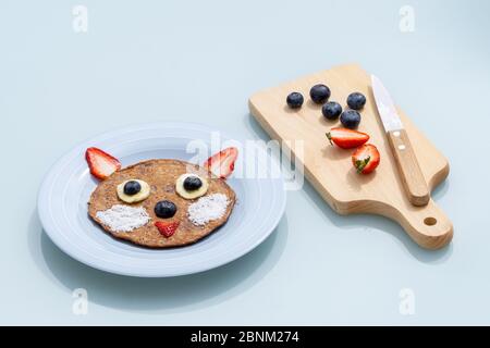 Kreativer Haferbrei Pfannkuchen für Kinder. Katze oder Bär in einer Platte auf blauem Hintergrund isoliert. Schneidebrett mit Früchten in der Nähe. Gesundes vegetarisches Frühstück, Mittagessen oder Snack. Kopierbereich. Stockfoto