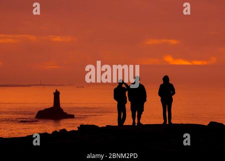 Touristen beobachten den Leuchtturm La Vieille in der Meerenge Raz de sein Silhouette gegen Sonnenuntergang an der Pointe du Raz, Plogoff, Finistere, Bretagne, F Stockfoto