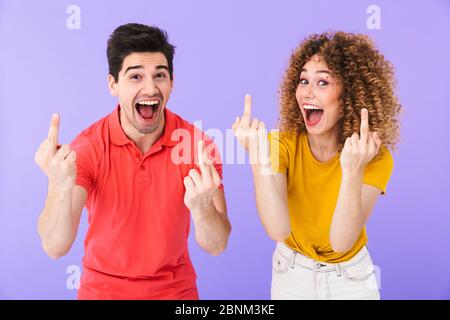 Porträt von optimistischen kaukasischen Menschen Mann und Frau in Grundkleidung lächelnd und zeigt Mittelfinger an der Kamera isoliert über violettem Hintergrund Stockfoto