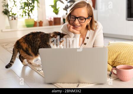 Freiberufliche Frau liegt im Wohnzimmer auf dem Teppich, Webinar ansehen. Frau mit ihrer Katze im Gespräch durch Video-Chat mit ihrer Familie, die in Isolation sind Dur Stockfoto