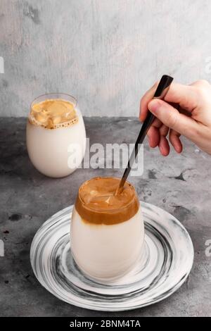 Beliebte Getränk koreanischen Dalgon Kaffee. Der lange Löffel mischt Schlagschaum aus löslichem Kaffee, Zucker und heißem Wasser mit Milch in Glas auf grauem Hintergrund Stockfoto