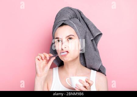 Unzufrieden und schläfrig junge Mädchen trinkt Kaffee und putzt ihre Zähne gleichzeitig auf rosa Hintergrund. Konzept Frühaufgänge, Morgenroutine Stockfoto