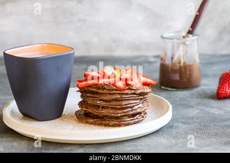 Schokolade Haferflocken Pfannkuchen Stapel mit Schokolade Sahne und Erdbeeren auf einem Teller. Tasse Kakao in der Nähe. Süßes Frühstück, Snack oder Dessert. Grauer Hintergrund. Nahaufnahme. Stockfoto