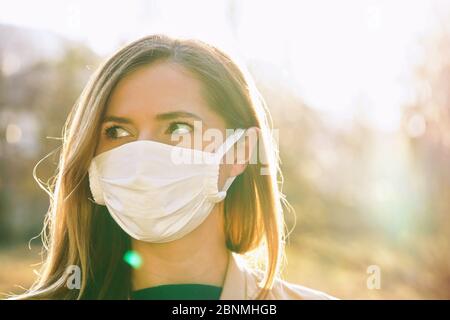 Junge Frau trägt weiße Baumwolle Virus Mund Nase Maske, schöne Hintergrundbeleuchtung Sonne Bokeh im Hintergrund, Nahaufnahme Gesicht Porträt Stockfoto