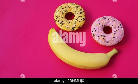 Zwei Donuts und eine Banane liegen auf der rosafarbenen Oberfläche und bilden eine Abstraktion des Emoticons - Lächeln. Vom oberen Punkt genommen Stockfoto