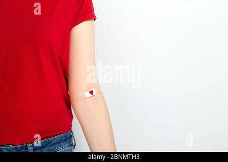 Blutspenderin. Junges Mädchen in rotem T-Shirt handtaped mit Patch mit rotem Herz nach dem Geben Blut auf grauem Hintergrund. Kopierbereich Stockfoto
