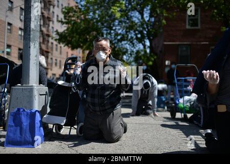 New York City, USA. Mai 2020. Ein Mann, der wegen der COVID-19-Pandemie eine Gesichtsmaske trägt, kniet auf dem Bürgersteig nieder, während er außerhalb der römisch-katholischen Kirche der Saint Bartholomew Parish bei Gottesdiensten über einen Lautsprecher betet, während die Menschen Nahrungsmittelhilfe erhalten, im Stadtteil Elmhurst des New Yorker Stadtbezirks Queens, NY, 15. Mai 2020. Stockfoto