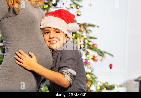 Porträt von Mutter und kleiner Junge trägt Santa Hut Umarmung Bauch Mutter in der Nähe Weihnachtsbaum Stockfoto