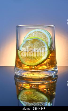 Ein Glas Whisky mit Eis und Zitrone auf blauem Hintergrund mit orangefarbener Hintergrundbeleuchtung und Reflektion. Stockfoto