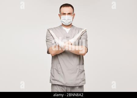 Männlicher Arzt in Uniform, Schutzmaske und Brille mit KEINER oder STOP-Geste, isoliert auf beigem Hintergrund. Konzept für Gesundheitswesen und Notfall Stockfoto