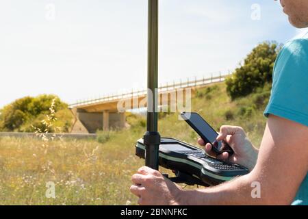 Landvermesser arbeitet mit einem GPS-Instrument, prüft Daten online mit seinem Smartphone. Außendienst Stockfoto