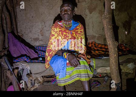 Ältere Maasai Frau in ihrem traditionellen Schlammhaus sitzt auf dem Bett ihres Mannes Stockfoto